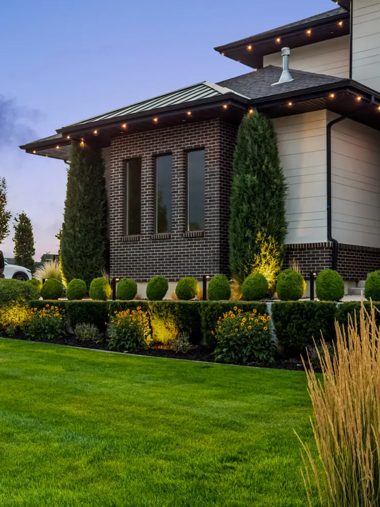 Cypress columnar topiaries and boxwood spheres accent lawn with warm uplighting along brick exterior.