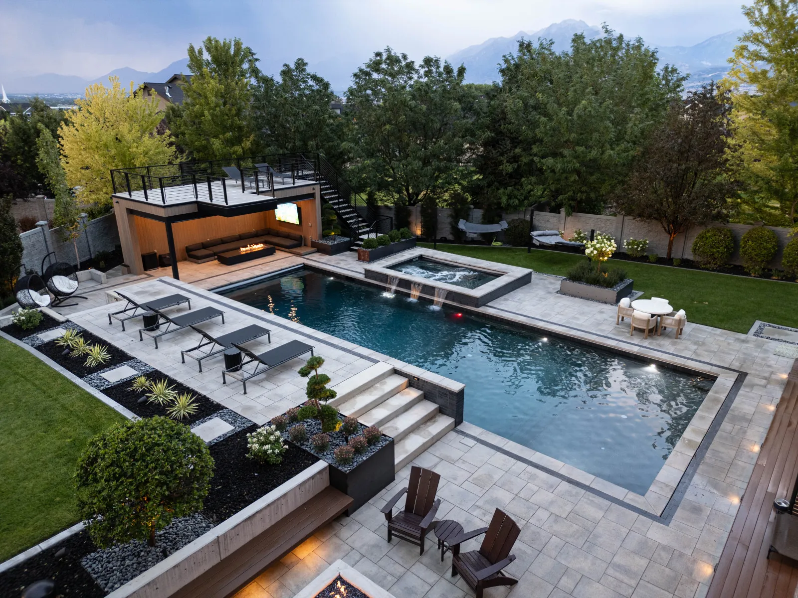 Infinity pool and paver patio with loungers, modern fire pit pavilion, landscaping, and mountain backdrop.